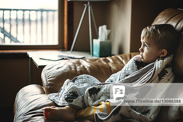 Nachdenklicher  in eine Decke gehüllter Junge  der sich zu Hause auf dem Sofa entspannt