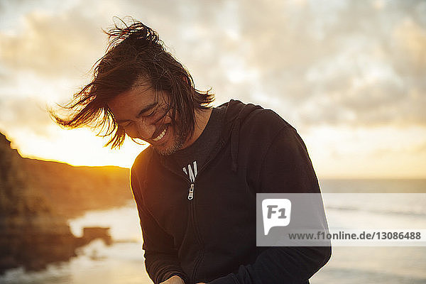 Happy man smiling while standing at beach against cloudy sky during sunset