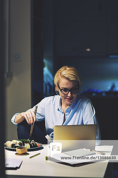 Businesswoman looking having food while using laptop computer at desk in home office