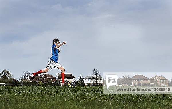 Side view of boy kicking soccer ball while playing on grassy field against sky