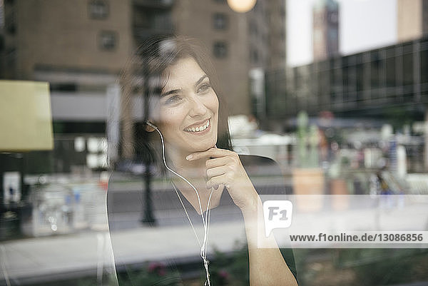 Happy woman listening music while looking through window in cafe seen through glass
