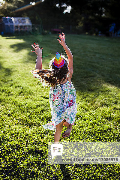 Fröhliches Mädchen mit Partyhut springt auf Grasfeld im Hinterhof