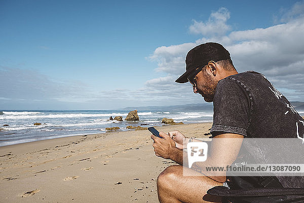 Side view of man using mobile phone while sitting at beach against blue sky during sunny day