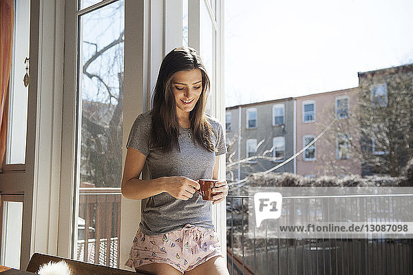 Frontansicht einer lächelnden Frau mit Kaffeetasse am Fenster stehend