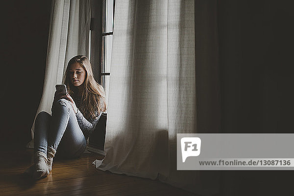 Teenager-Mädchen in voller Länge benutzt ein Smartphone  während sie zu Hause am Fenster sitzt