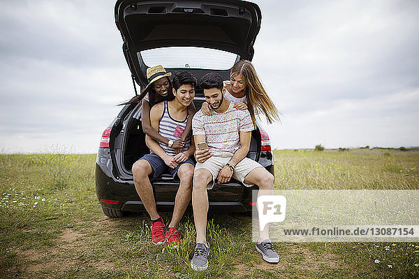 Happy friends using smart phone while sitting in car trunk on field against sky