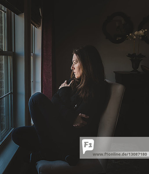 Nachdenkliche Frau schaut durch das Fenster  während sie zu Hause auf einem Stuhl sitzt