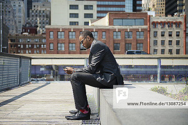 Seitenansicht eines Geschäftsmannes  der ein Smartphone benutzt  während er auf einer Stützmauer sitzt