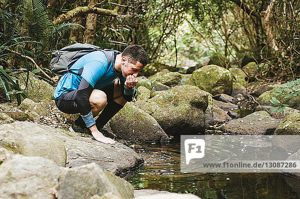Male hiker drinking water from lake while crouching on rocks in forest