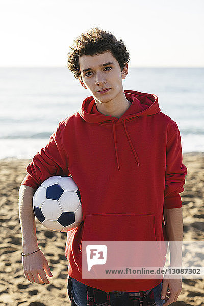 Portrait of confident teenage boy holding soccer ball while standing at beach against sea