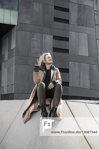 Glückliche Frau sitzt auf Stützmauer außerhalb des grauen Gebäudes