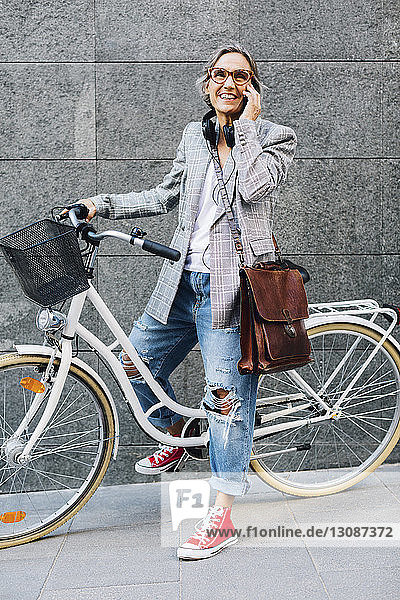 Happy woman talking on smart phone with bicycle against wall on footpath
