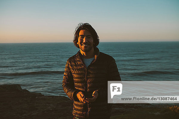 Porträt eines glücklichen Mannes  der ein Smartphone in der Hand hält  während er bei Sonnenuntergang am Meer steht