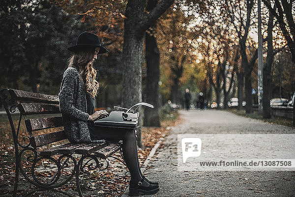 Seitenansicht einer Schriftstellerin mit Schreibmaschine  die im Herbst auf einer Parkbank sitzt