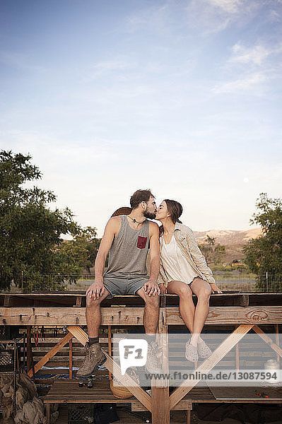 Liebevolles Bauernpärchen küsst sich  während es auf einer Holzkonstruktion auf dem Bauernhof gegen den Himmel sitzt