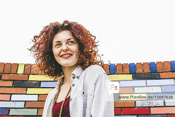 Low angle view of woman looking away while standing by colorful brick wall against clear sky