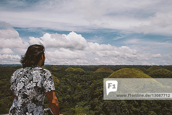 Rear view of man looking at Chocolate Hills while standing by railing against cloudy sky
