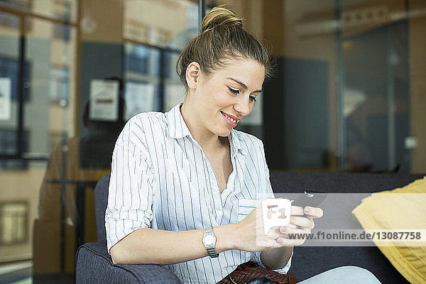 Lächelnde Geschäftsfrau benutzt Mobiltelefon  während sie im Büro auf dem Sofa sitzt