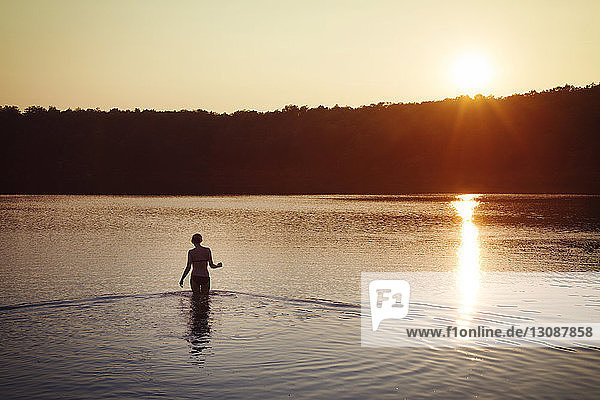 Rückansicht einer Frau im Bikini  die bei Sonnenuntergang im See steht