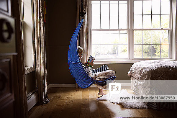 Boy reading book while sitting on swing in bedroom