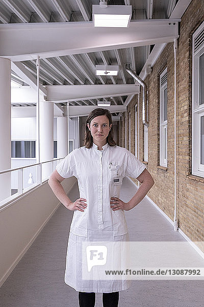 Portrait of confident female doctor with hands on hip standing in hospital corridor