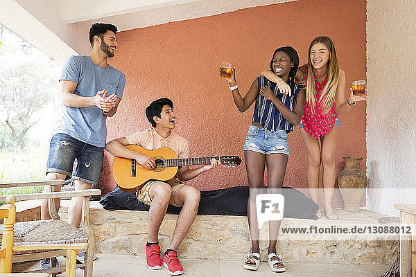 Cheerful women holding iced tea glasses while male friend playing guitar at yard