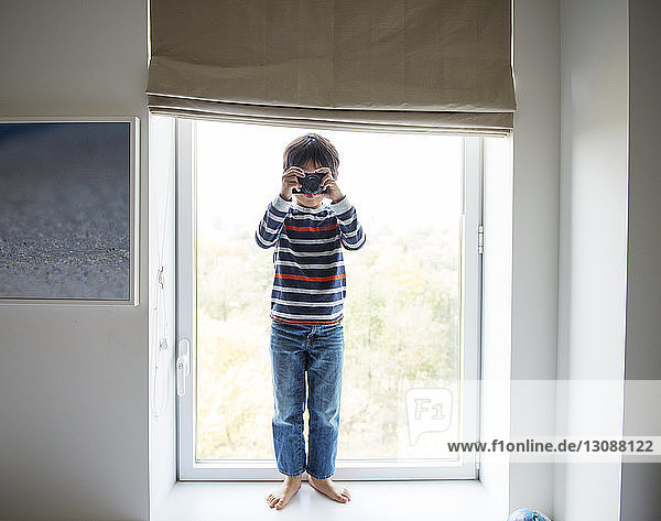 Fröhlicher Junge spielt zu Hause mit der Kamera auf dem Fensterbrett
