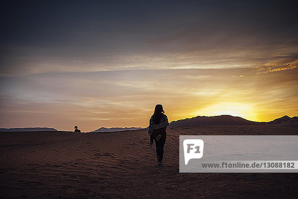 Rückansicht einer auf Sand stehenden Frau in der Wüste vor bewölktem Himmel bei Sonnenuntergang