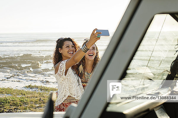Happy female friends taking selfie while standing by off-road vehicle at beach