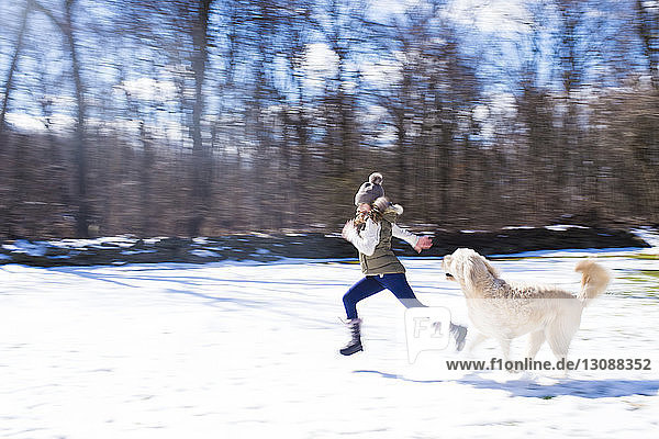 Side view of girl with dog running on snow covered field against bare trees