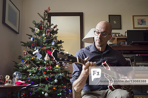 Mann spielt Gitarre  während er zu Hause am Weihnachtsbaum sitzt