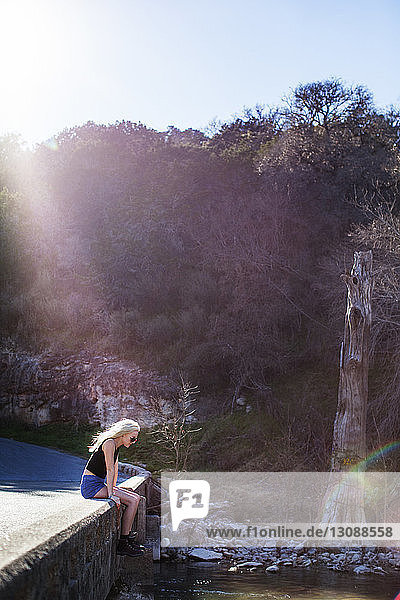 Seitenansicht einer jungen Frau  die an einem sonnigen Tag auf einer Stützmauer am Fluss sitzt