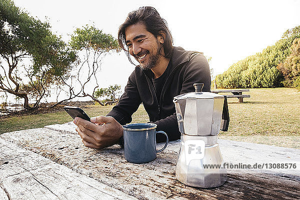 Glücklicher Mann mit Kaffee auf Holztisch  der mit seinem Handy gegen den Himmel sitzt