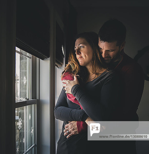 Couple embracing while standing by window at home