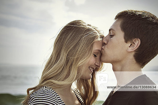 Side view of man kissing girlfriend's forehead standing at beach