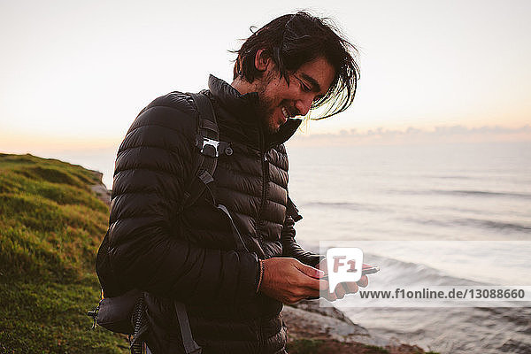 Glücklicher männlicher Wanderer  der ein Smartphone benutzt  während er auf einem Hügel am Meer steht