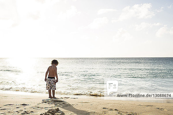 Rückansicht eines Jungen ohne Hemd  der am Strand gegen den Himmel an einem sonnigen Tag steht