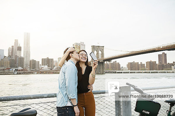 Man kissing happy woman photographing against East river in city
