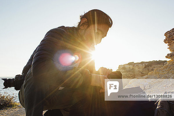 Low angle view of man with backpack crouching at beach against clear sky during sunset