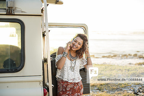 Happy woman talking on mobile phone by off-road vehicle at beach