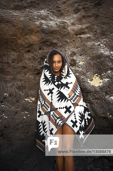 Portrait of teenager wrapped in blanket standing against rock