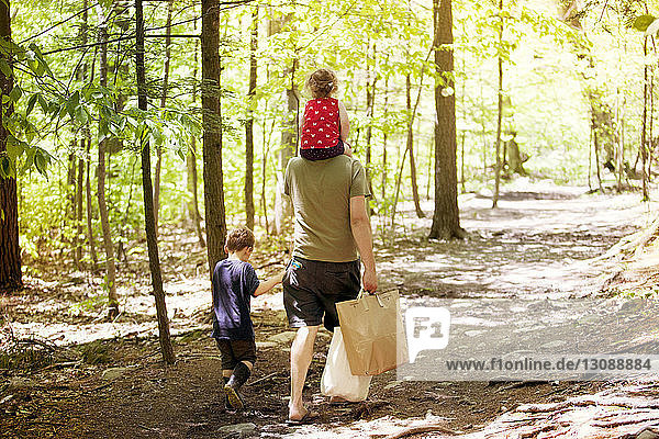 Rückansicht eines Vaters mit Taschen in der Hand  der mit seinem Sohn und seiner Tochter auf einem Feldweg inmitten des Waldes geht