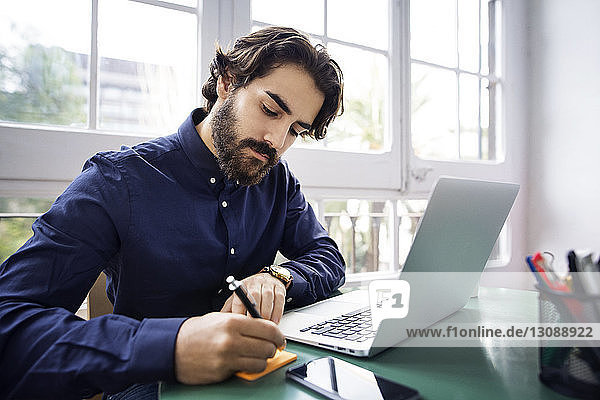 Businessman writing on adhesive note while sitting at table with laptop in office