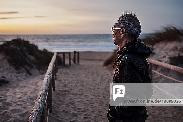Thoughtful man standing at beach against sky during sunset