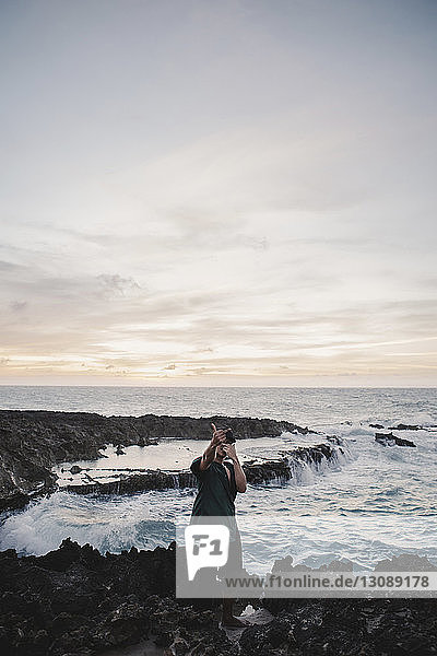 Mann zeigt obszöne Geste  während er auf Felsen am Meer steht