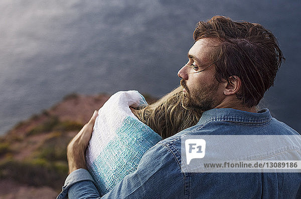 Rückansicht eines nachdenklichen Mannes  der eine Frau umarmt  während er am Meer sitzt