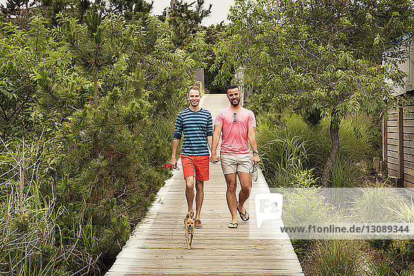 Full length of homosexual couple with Chihuahua on boardwalk amidst trees