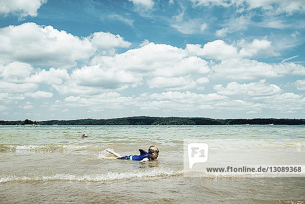 Boy wearing swimming goggles while lying in sea against cloudy sky