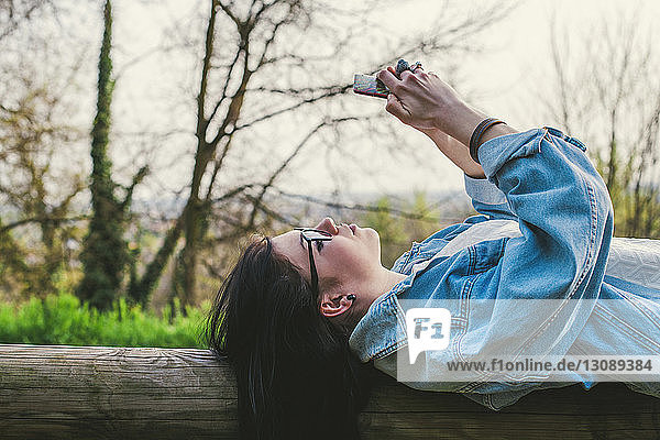 Seitenansicht einer Frau  die ein Mobiltelefon benutzt  während sie auf einem Holzgeländer gegen den Himmel im Park liegt