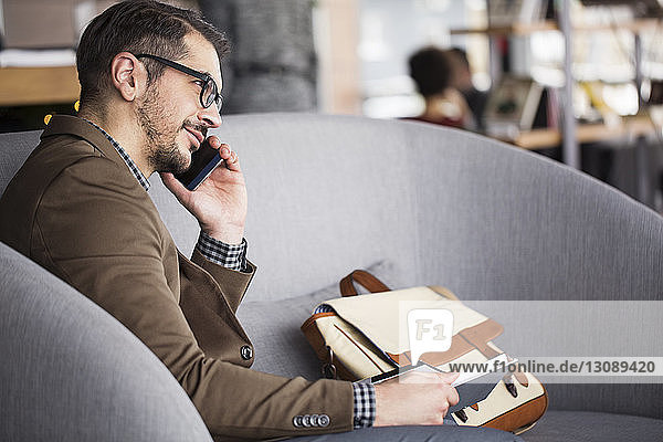 Lächelnder Geschäftsmann hält Tagebuch  während er im Restaurant am Smartphone telefoniert